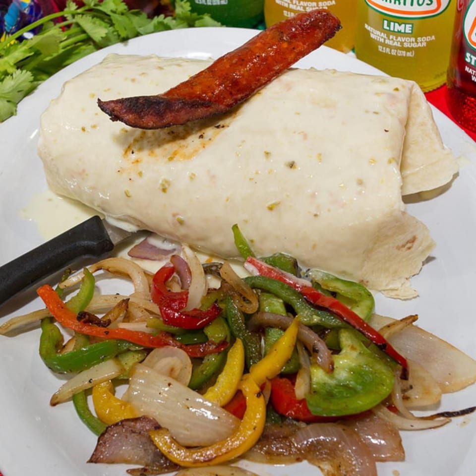 A large burrito topped with chorizo, served with grilled peppers and onions on a white plate.