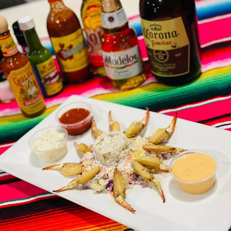 Crab claws and shredded crab meat with sauces, hot sauce bottles, and beer on a colorful Mexican blanket.