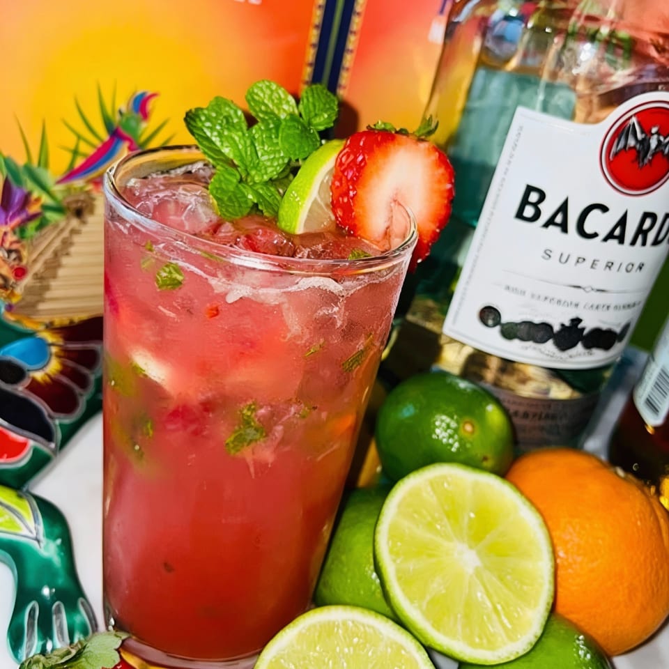 Strawberry mojito with mint and lime, Bacardi bottle, and colorful Mexican decor in the background.