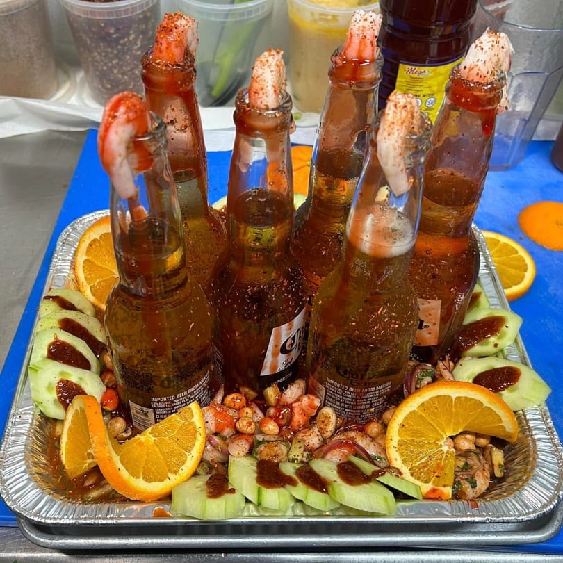 Six beer bottles with shrimp and chili-lime seasoning, surrounded by cucumber, orange slices, and ceviche.